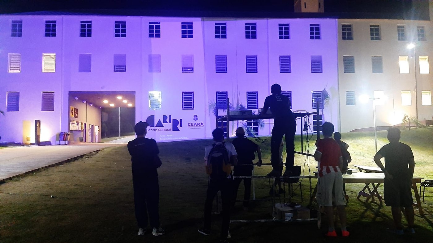 Fachada mapeada em roxo, andaime de projeção e turma reunida na grama