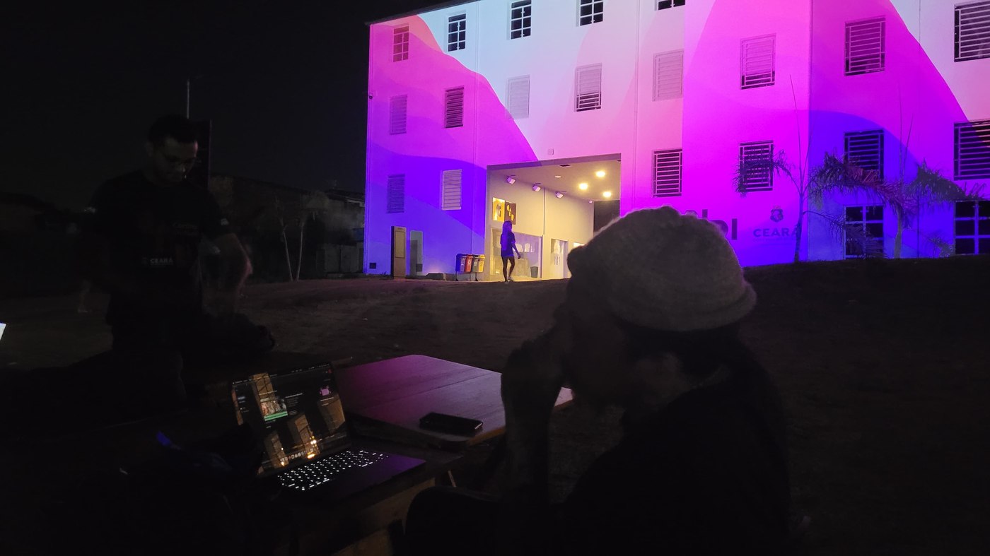 Conteúdo em gradiente rosa, roxo e azul projetado na fachada, notebook em primeiro plano