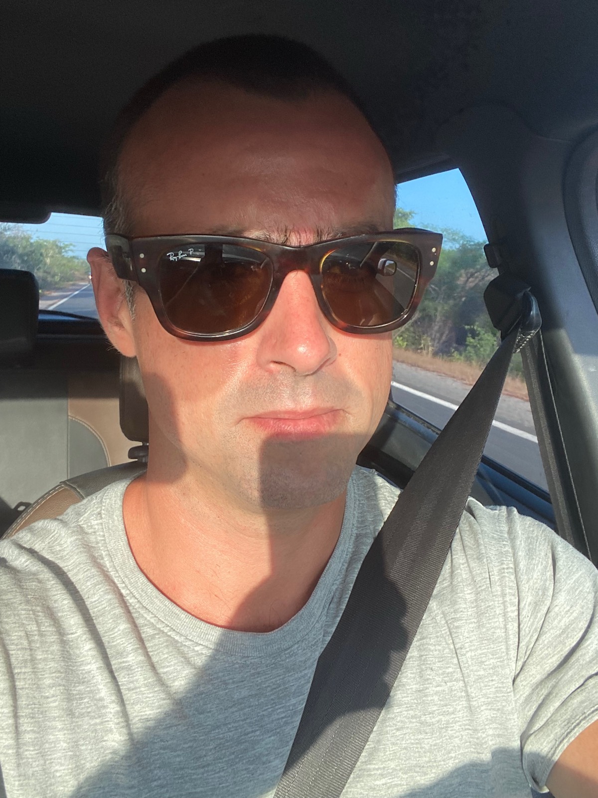 Selfie dirigindo, de óculos de sol, com a estrada refletida no vidro do carro