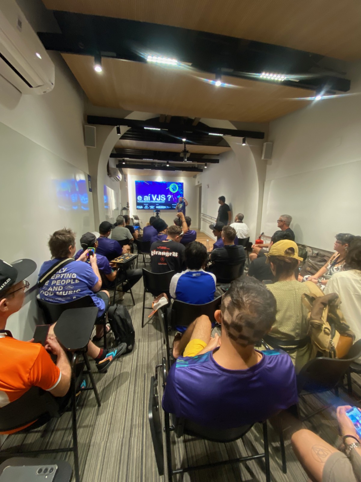 Sala cheia de VJs sentados assistindo a uma palestra, tela ao fundo perguntando 'E aí VJs, vocês estão preparados?'