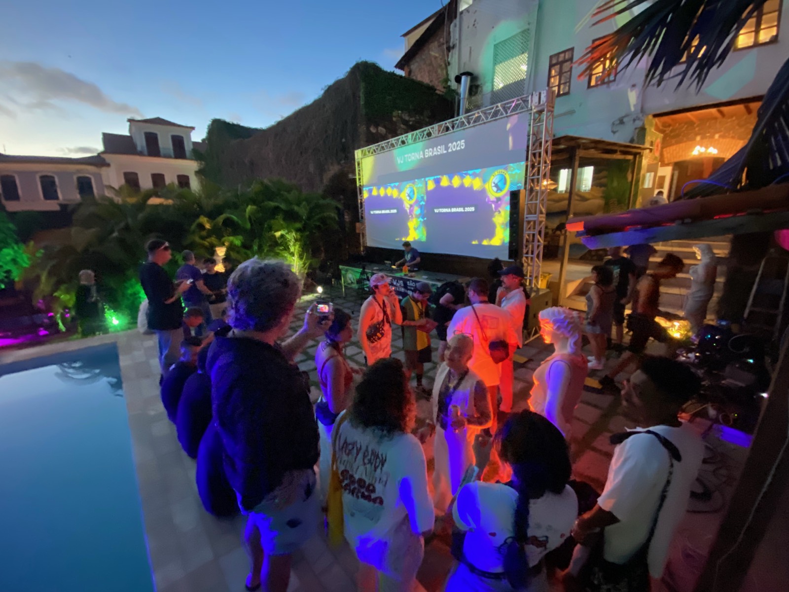 Pátio da pousada de noite, com piscina e tela grande projetando VJ Torna Brasil 2025