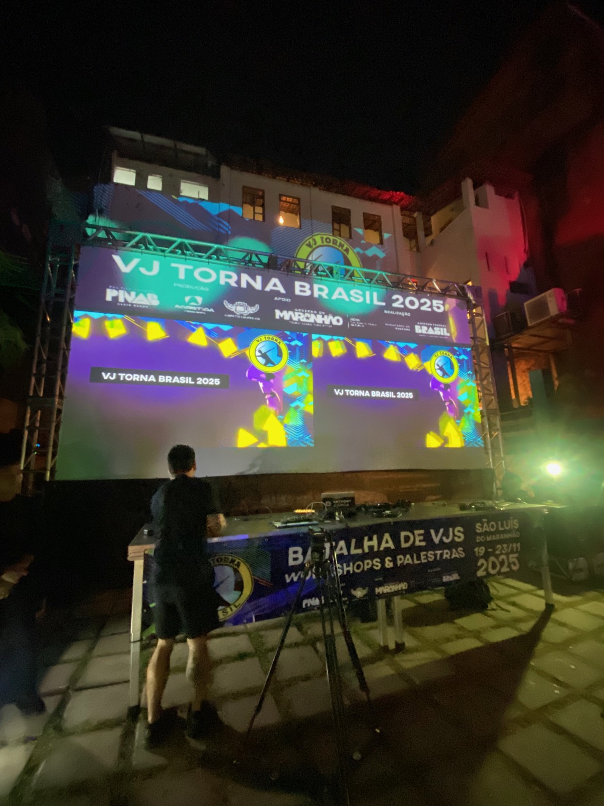 Tela grande vista de longe, com banner 'Batalha de VJs, Workshops & Palestras' e mesa de som/projeção em primeiro plano, à noite