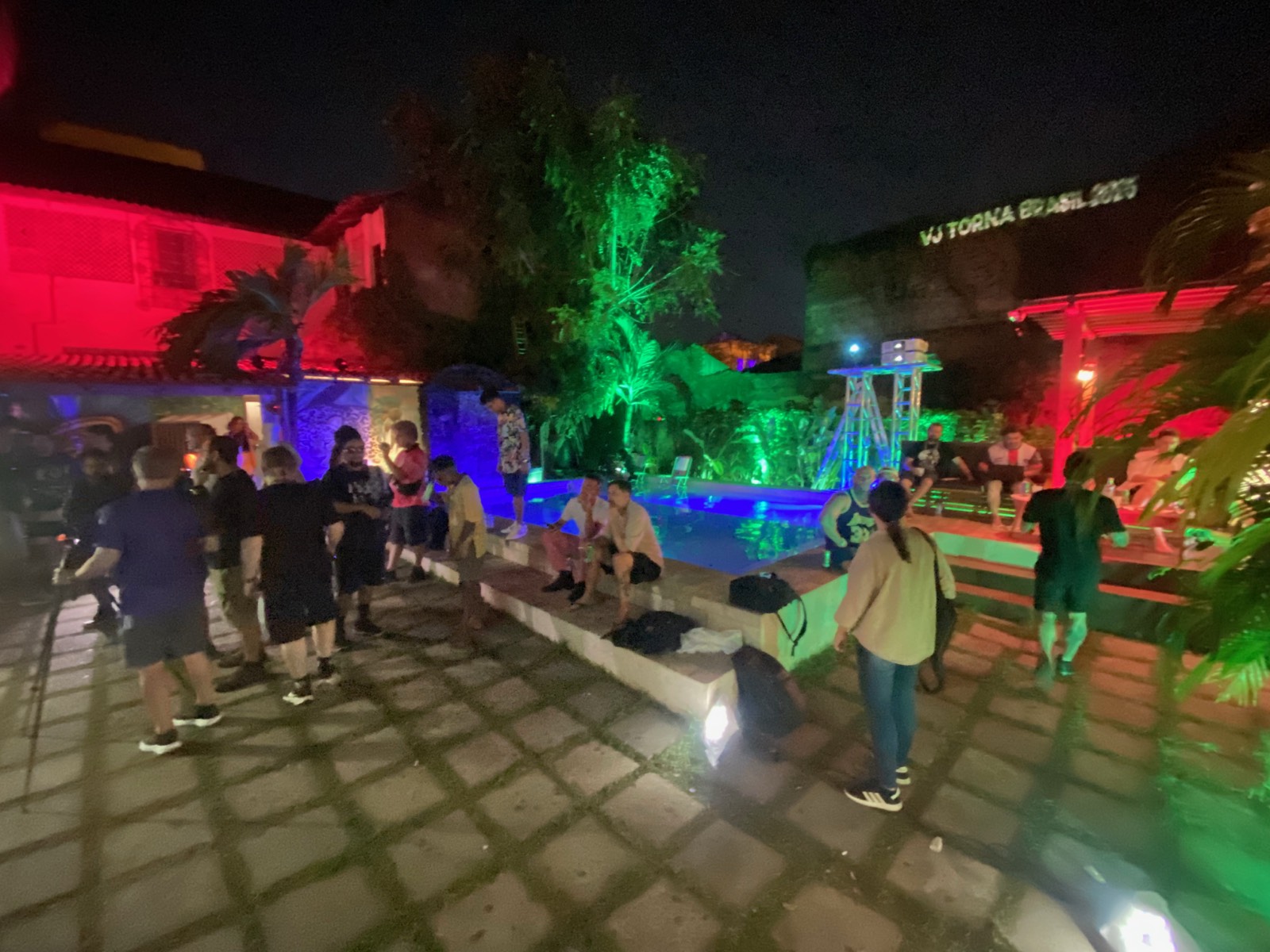 Pátio da pousada à noite, com luz verde e vermelha, piscina e convidados reunidos