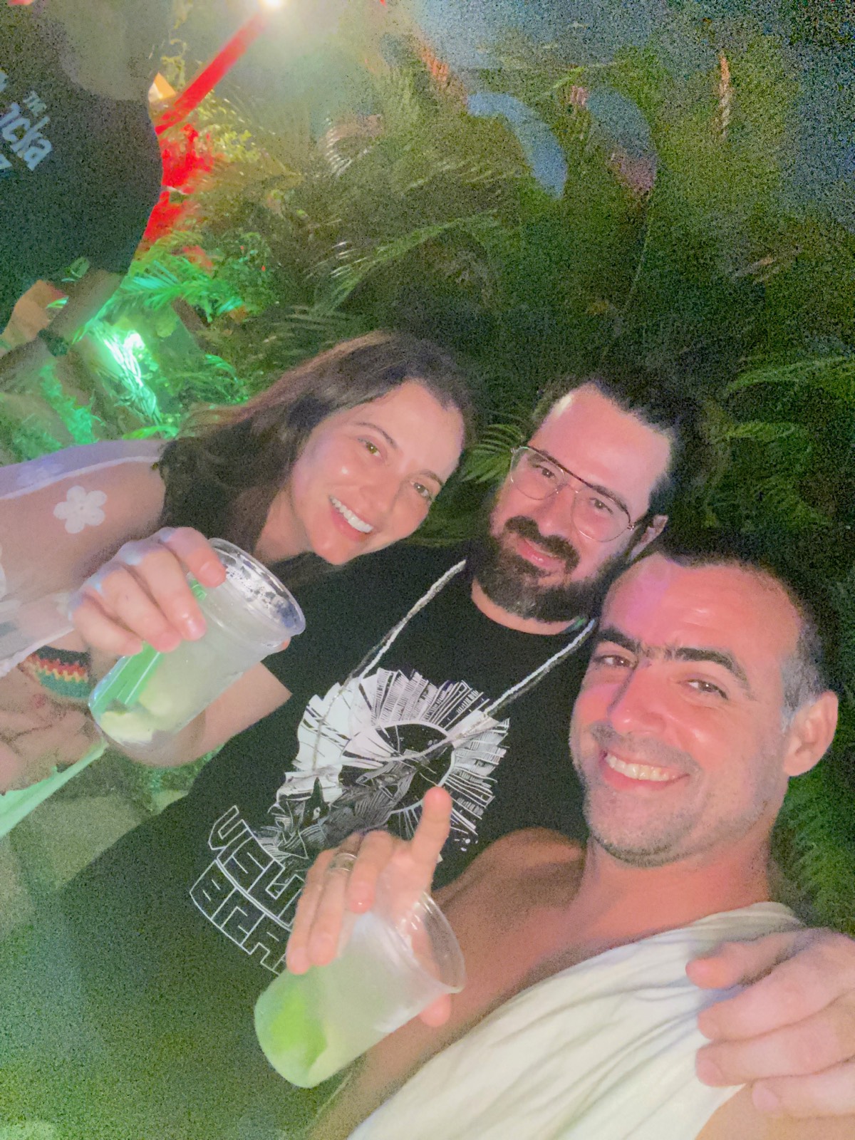 Selfie em trio de noite, com drinques na mão, vegetação ao fundo