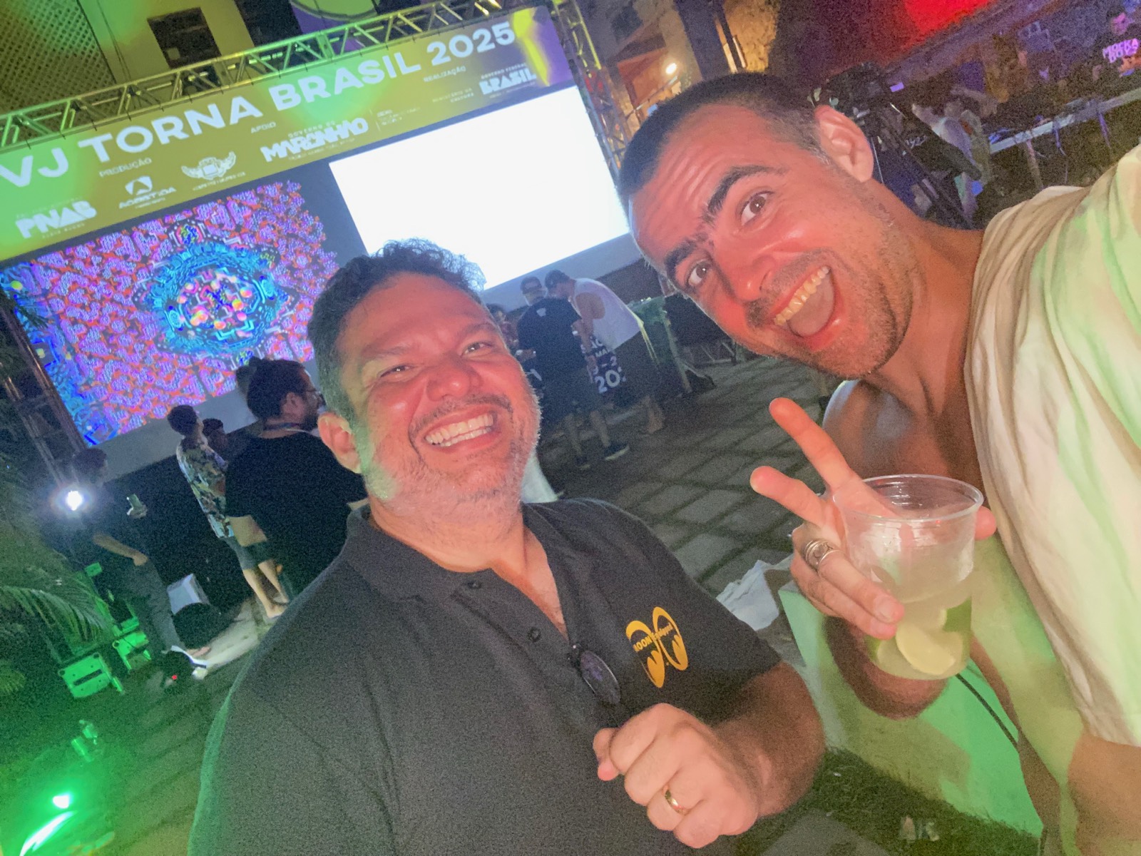 Selfie em dupla de noite, com arte generativa projetada na tela grande ao fundo