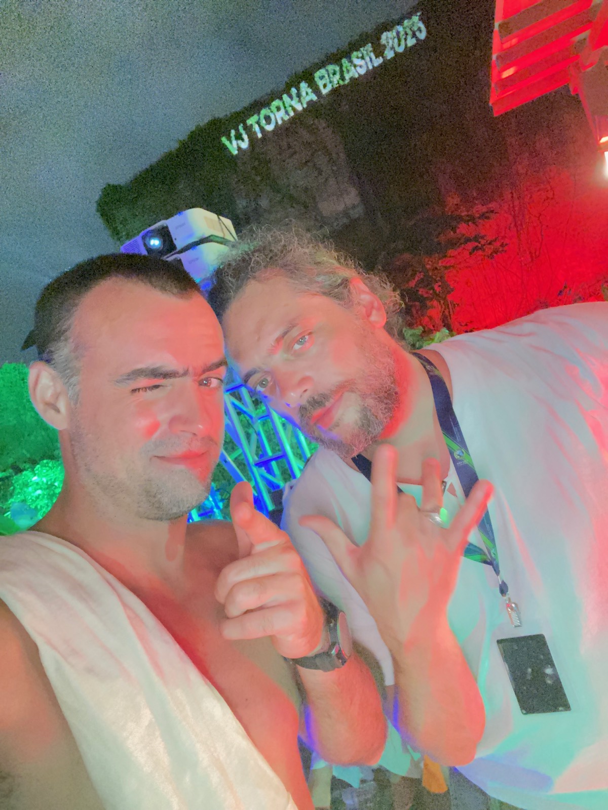 Selfie em dupla de noite, com o letreiro 'VJ Torna Brasil' iluminado ao fundo